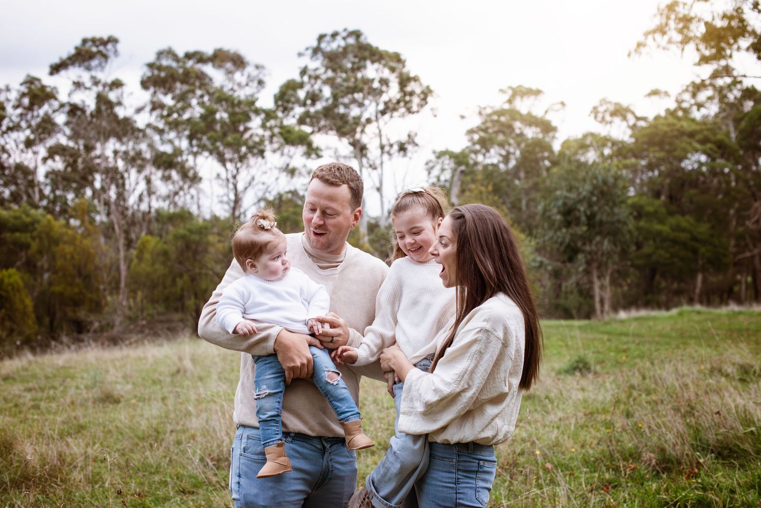 Happy family of four laughing together in a green meadow with tall eucalyptus trees in the background.