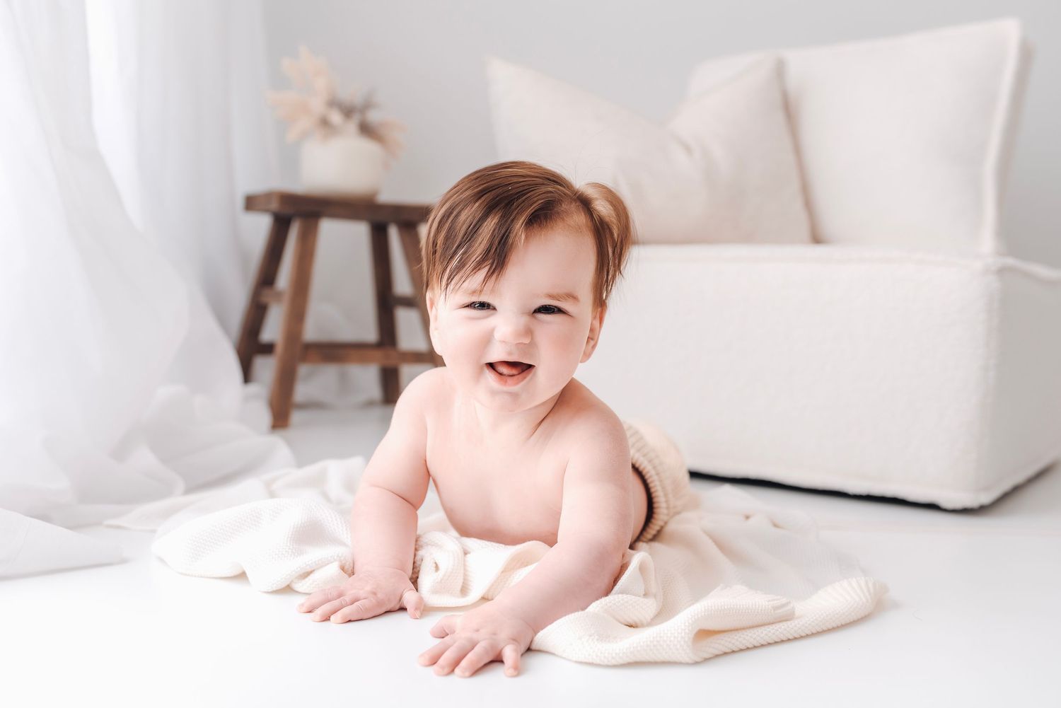 A laughing baby with dark hair sits on a white blanket in a bright minimalist white room.