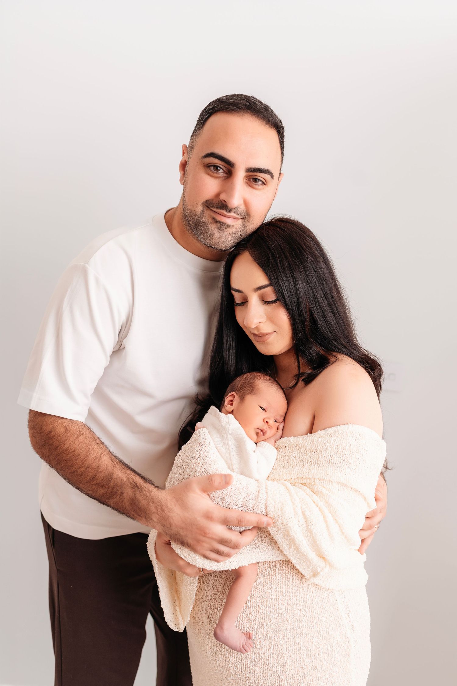 Parents in white outfits lovingly hold their sleeping newborn baby in a studio portrait.