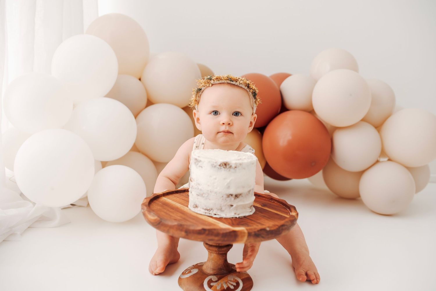 A sweet baby girl stands at a white frosted smash cake on a wooden stand with boho balloon backdrop.