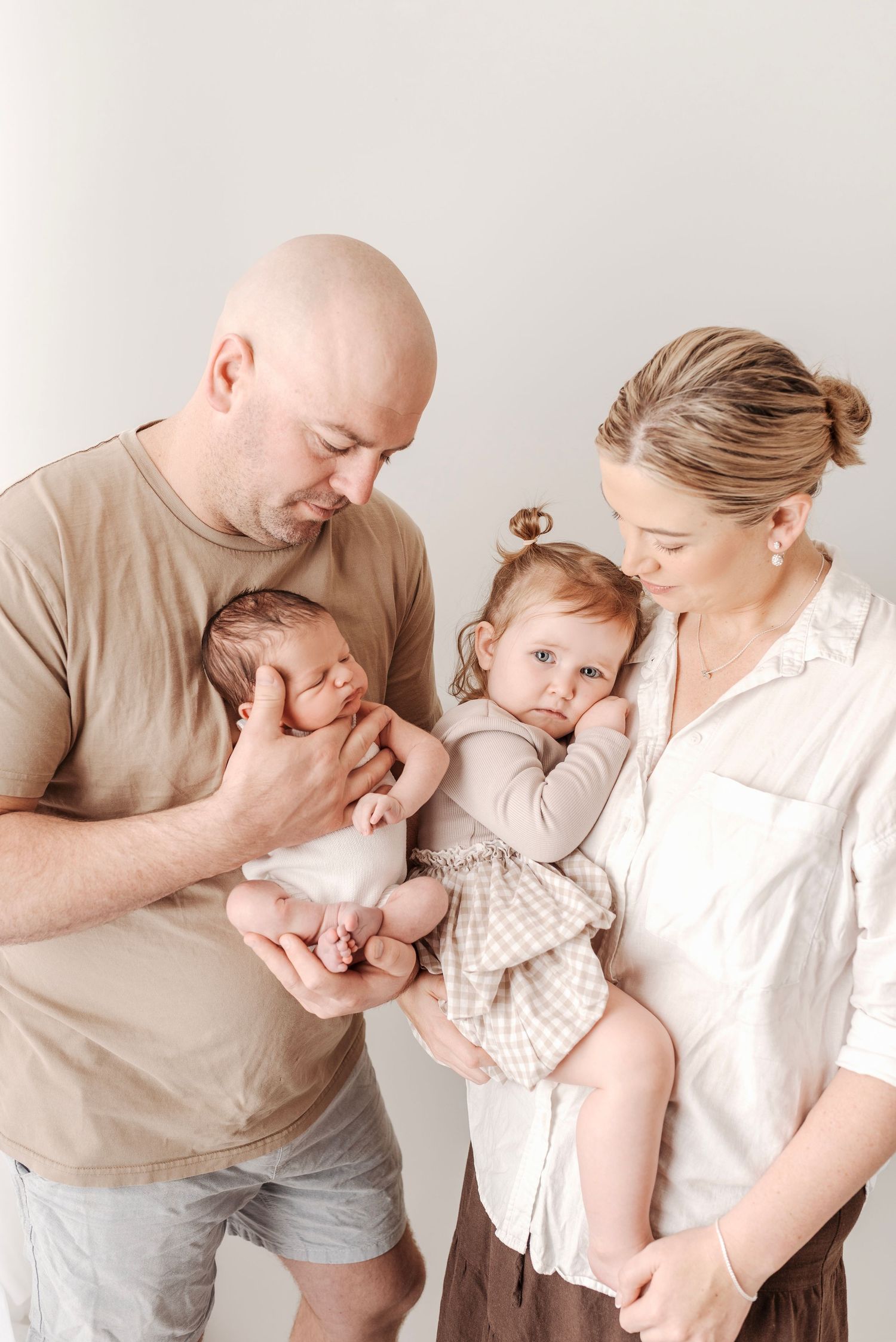 Bald father holds sleeping newborn while mother carries toddler daughter in a warm family portrait.