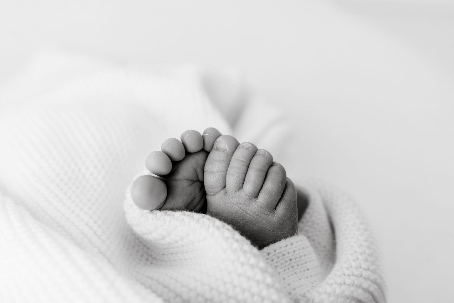 Black and white close-up photo of a newborn baby's tiny feet wrapped in a soft white knit blanket.