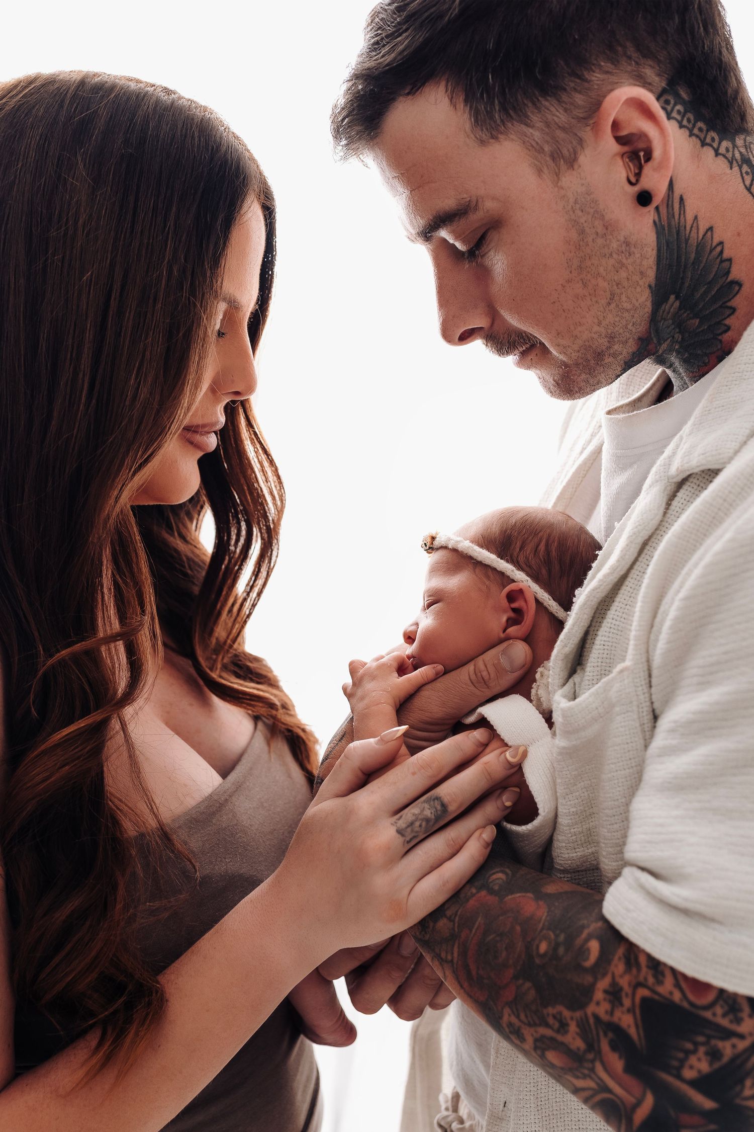 Tattooed parents gently cradle their newborn baby together in a tender close-up portrait.