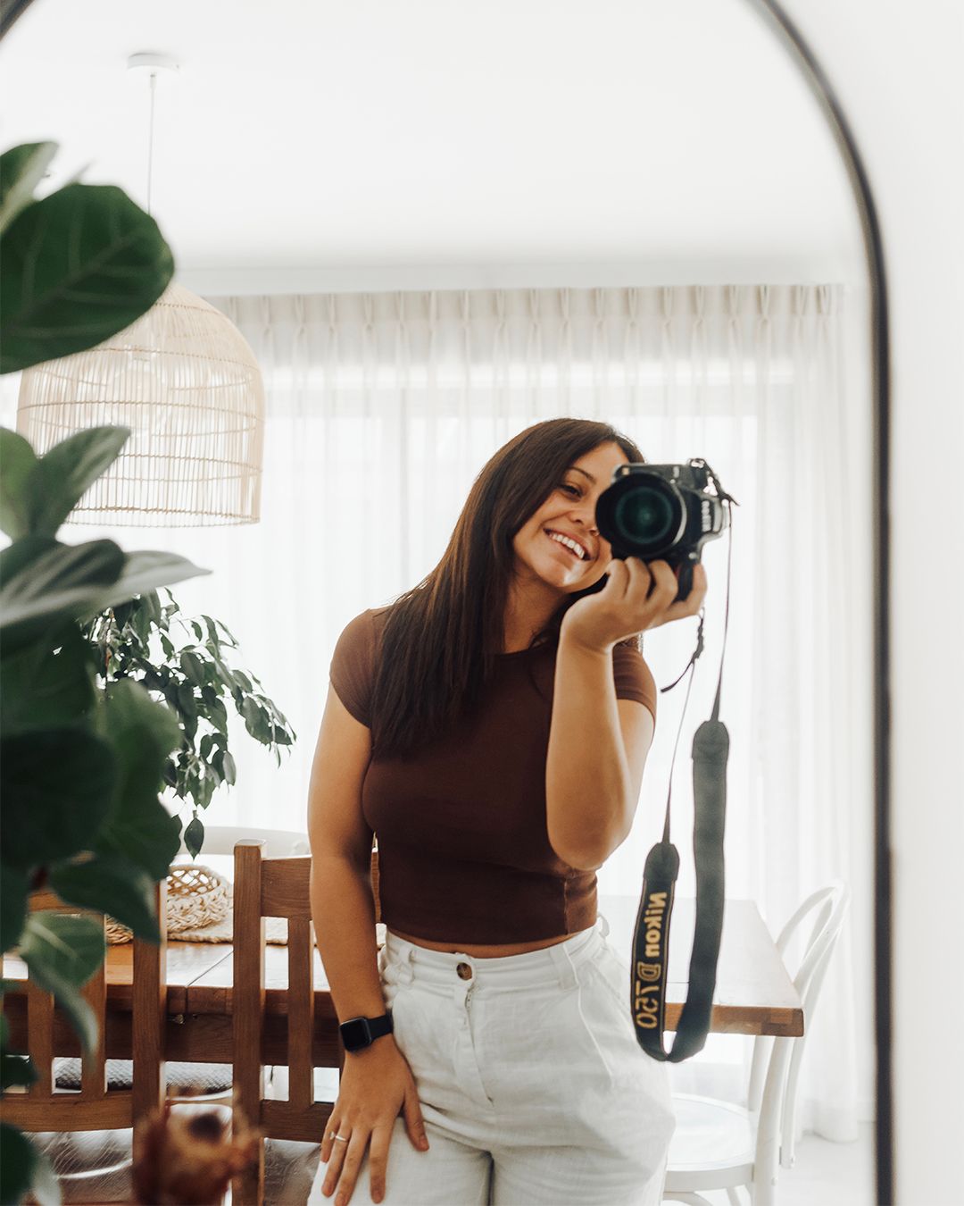 Young woman smiling while taking mirror selfie with DSLR camera in bright modern home.