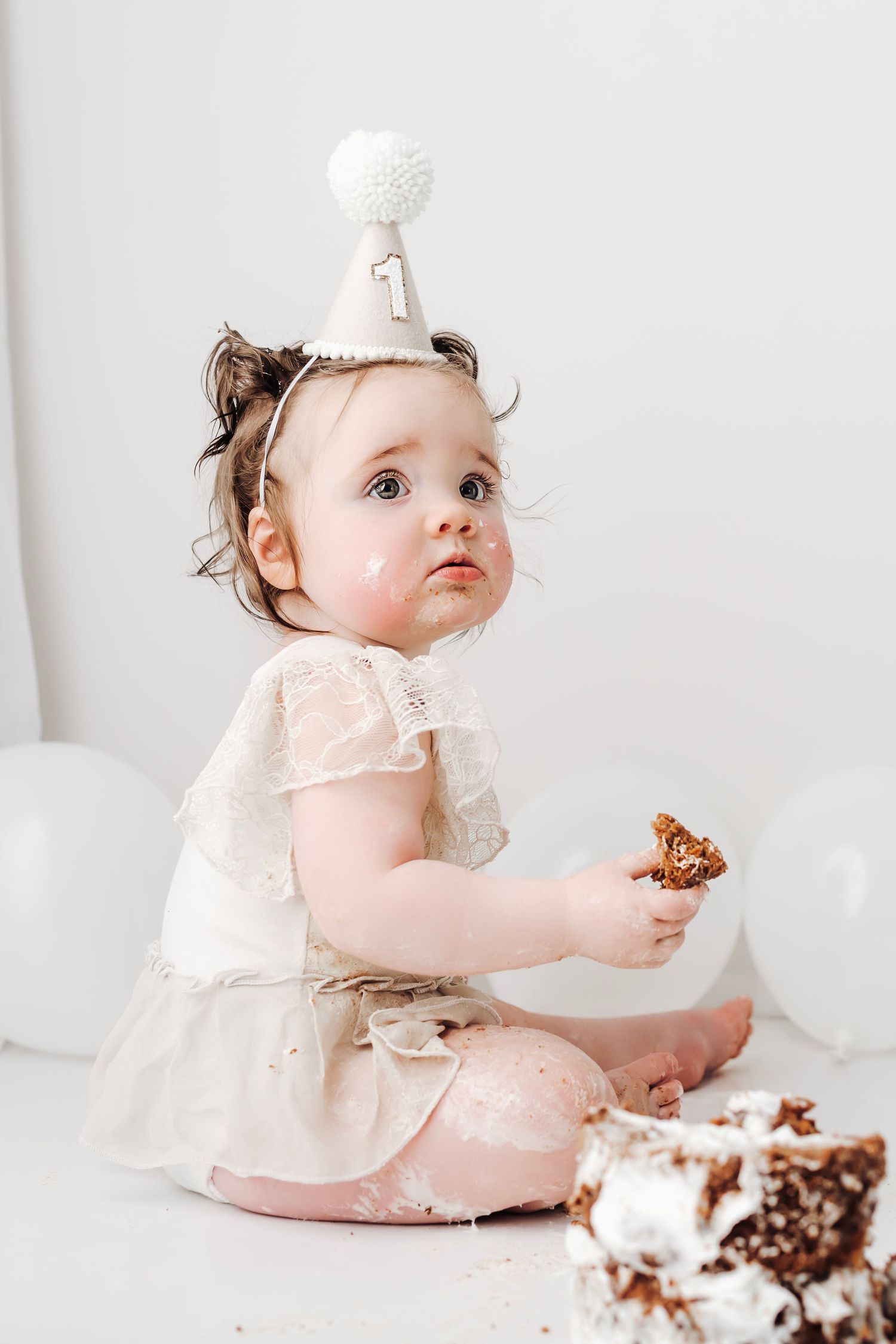 A baby girl in a white dress and party hat enjoys her first birthday cake smash, holding a piece of cake.