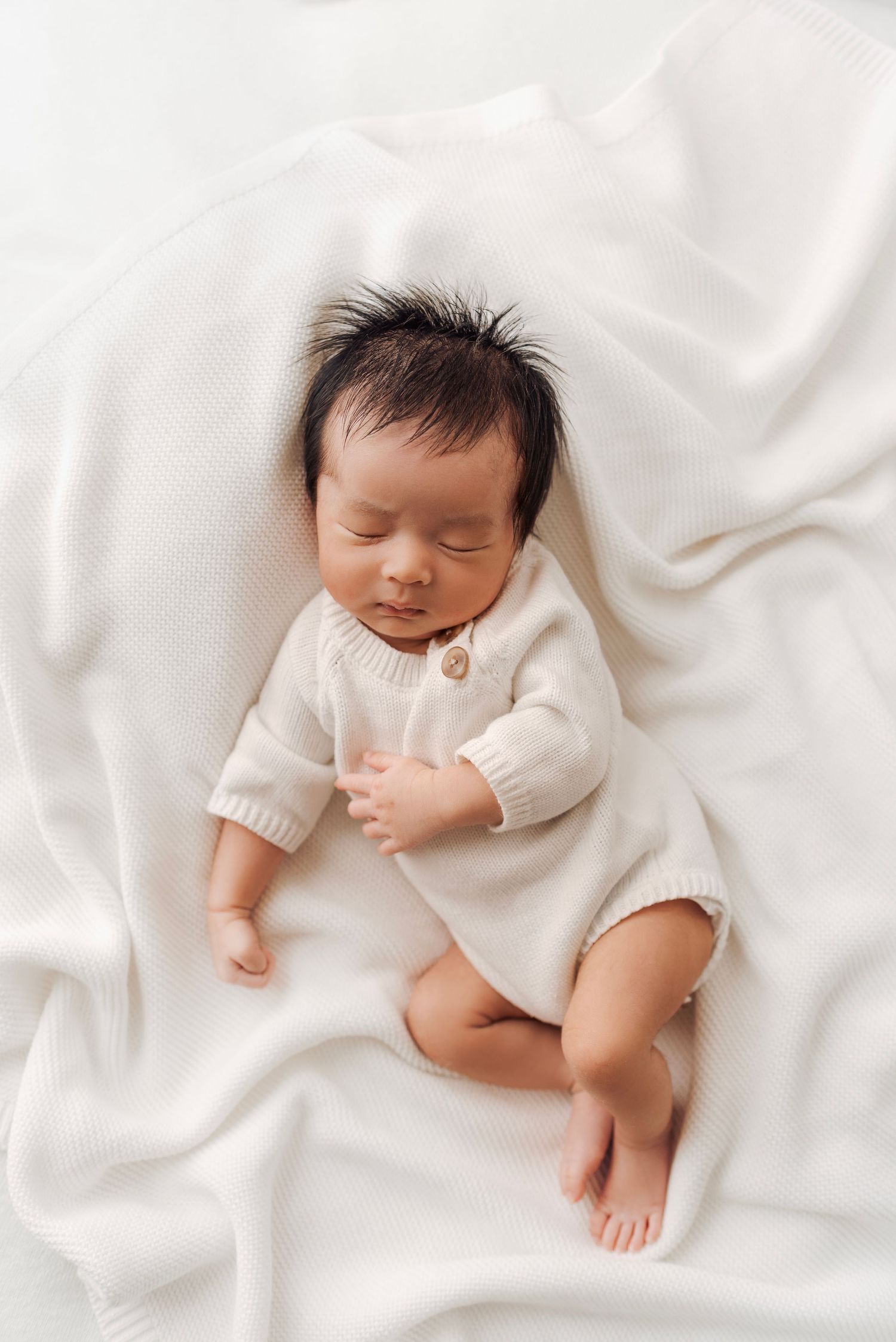 Newborn baby sleeping peacefully in cream knit romper on soft white blanket.