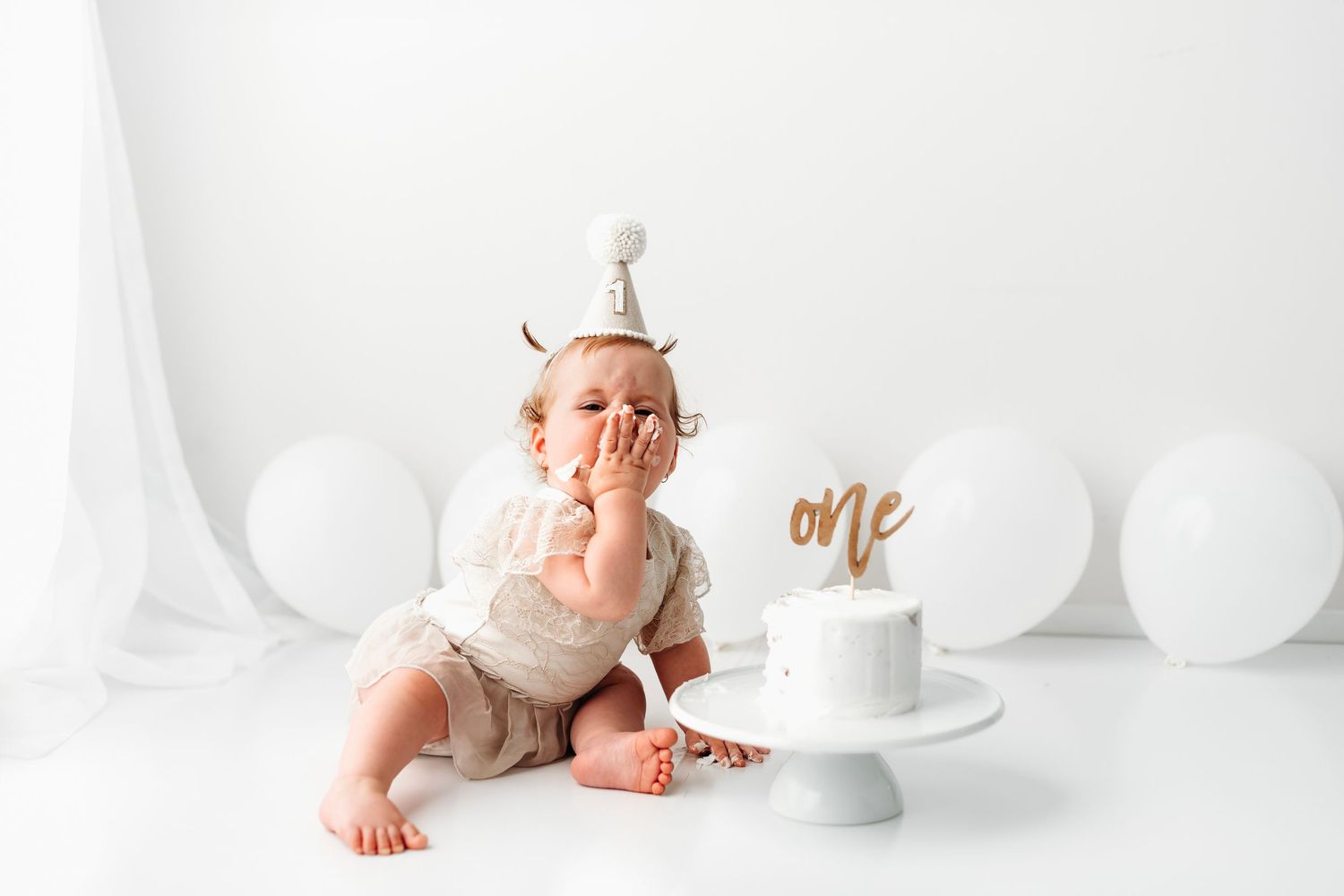 A baby in a white outfit and party hat covers their mouth while sitting near a cake with an 'one' topper.