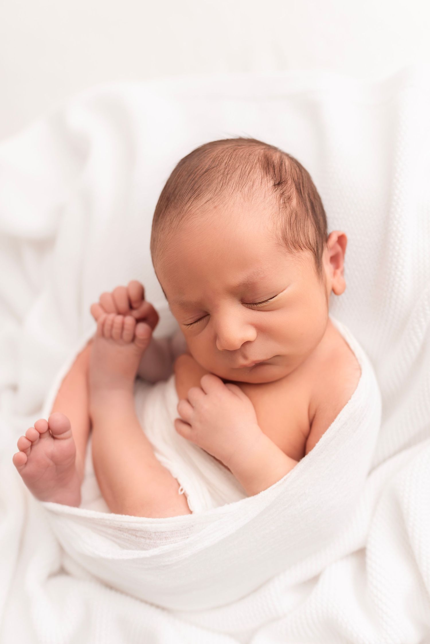Peaceful newborn baby wrapped in white swaddle blanket sleeping with tiny fist raised on soft white surface.
