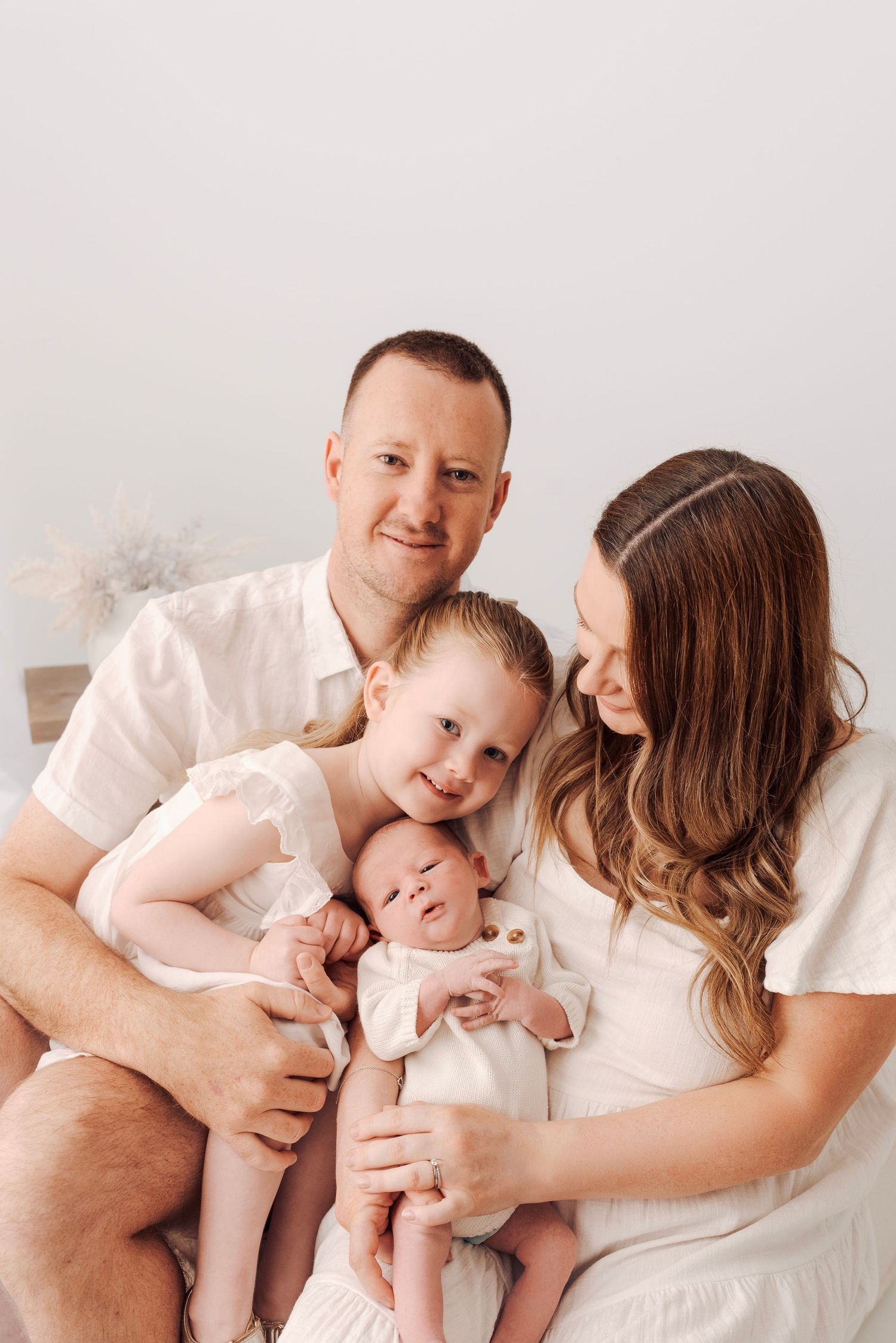 A happy family of four poses together, with parents holding a toddler and newborn baby, dressed in white outfits.