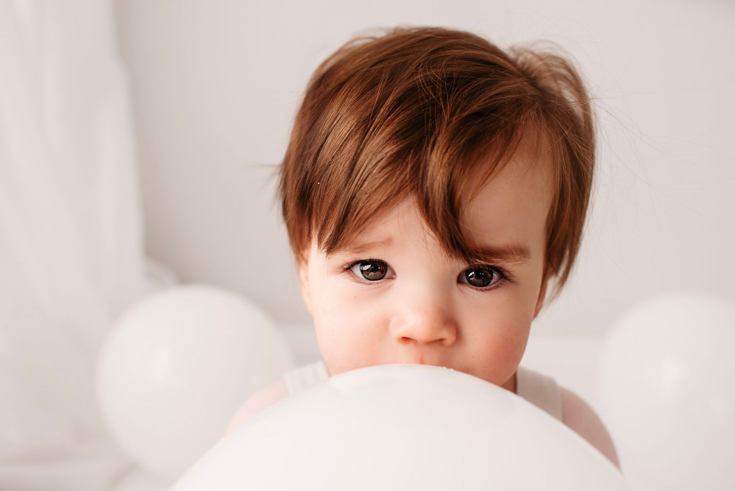 Adorable toddler with brown hair peeking over white balloon with curious expression in bright white setting.