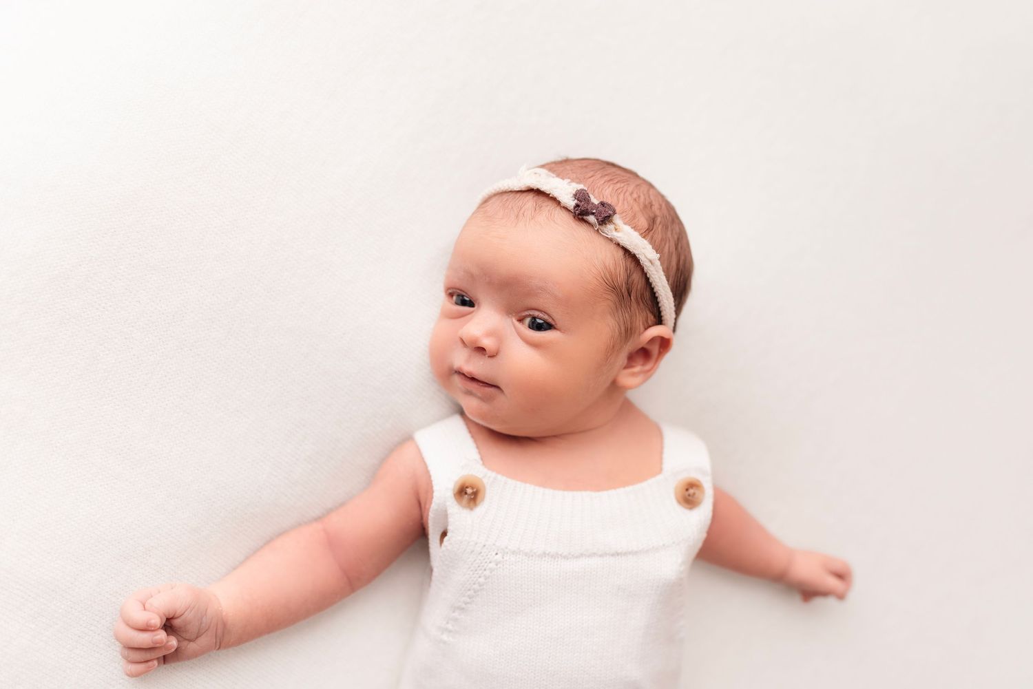 Newborn baby girl in white romper with floral headband lies on white backdrop with arms outstretched.
