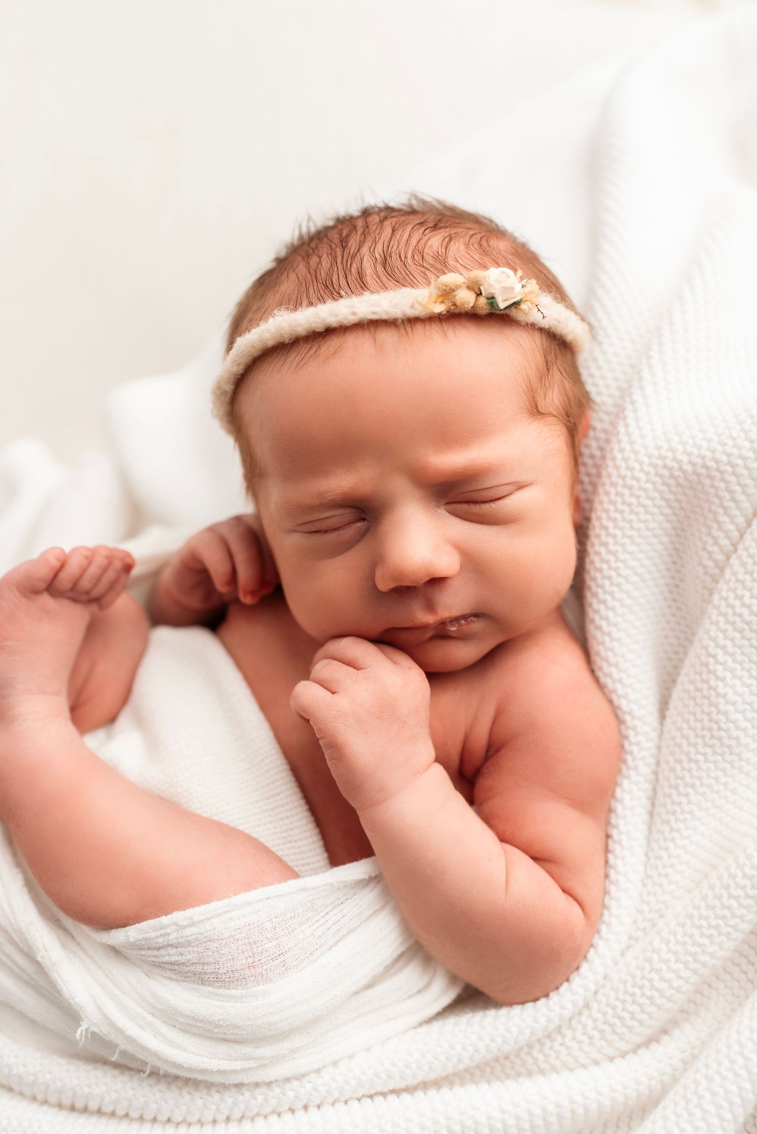 Peaceful newborn baby girl sleeping wrapped in white knit blanket wearing a delicate floral headband.