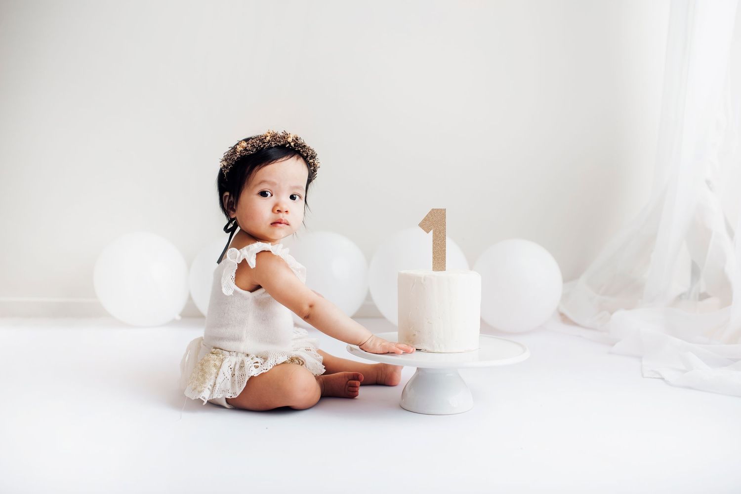 Adorable baby girl in white outfit celebrating first birthday with cake and balloon decorations.
