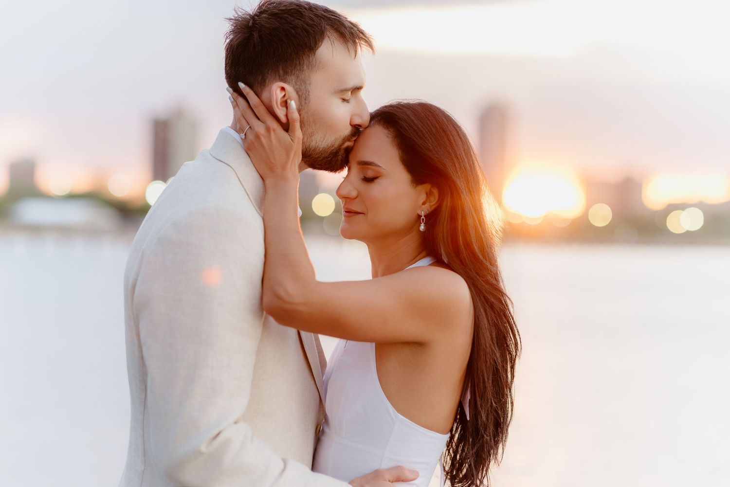 Groom kissing bride on forehead during golden sunset by the waterfront.