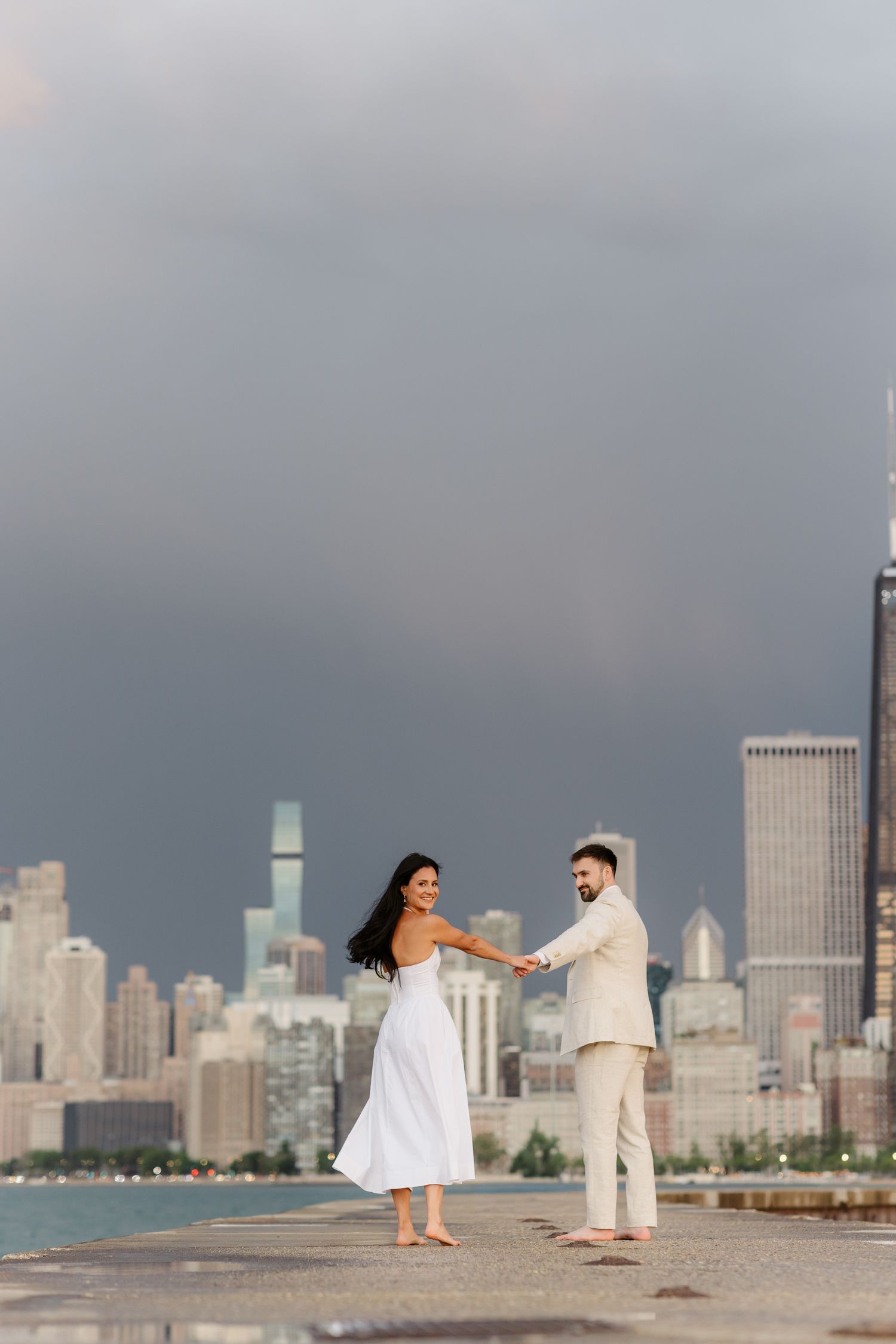Smiling couple dances barefoot on waterfront pier with dramatic cloudy Chicago skyline in background.