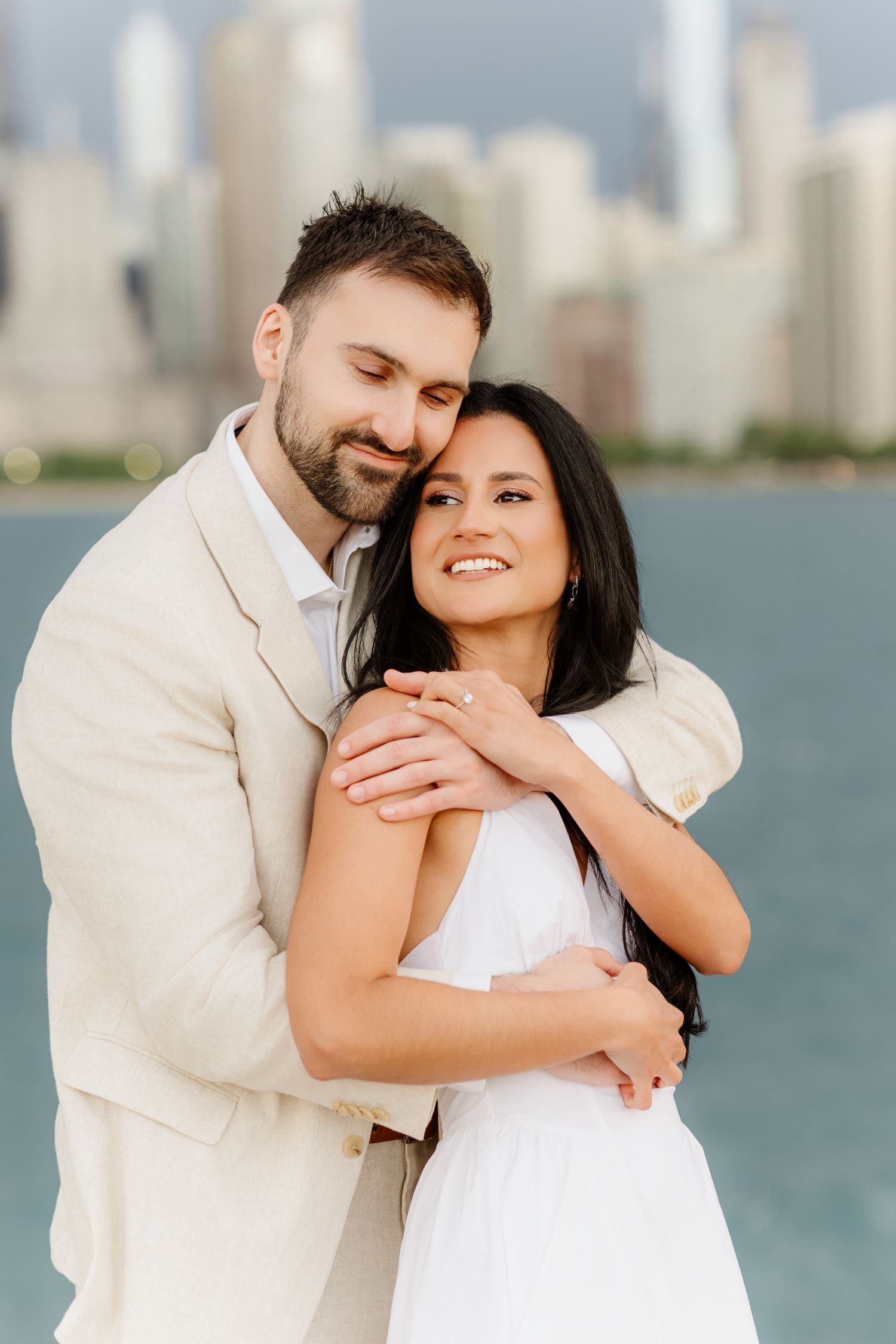 Romantic couple embracing near Chicago waterfront, man in tan suit nuzzling woman in white dress with skyline backdrop.