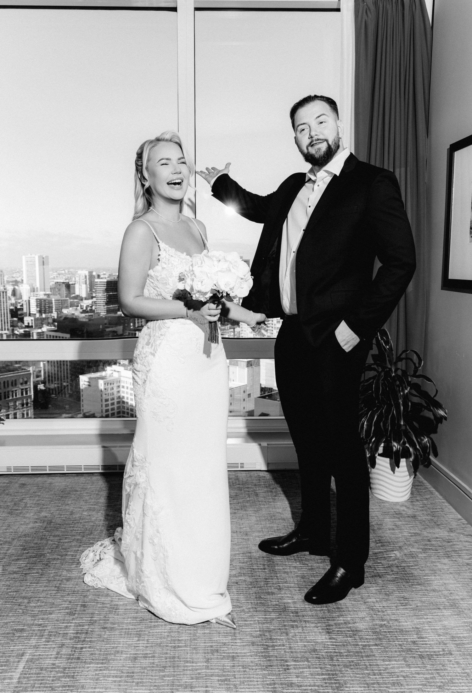 Black and white photo of laughing bride in lace gown and bearded groom in tuxedo posing playfully in high-rise hotel room.