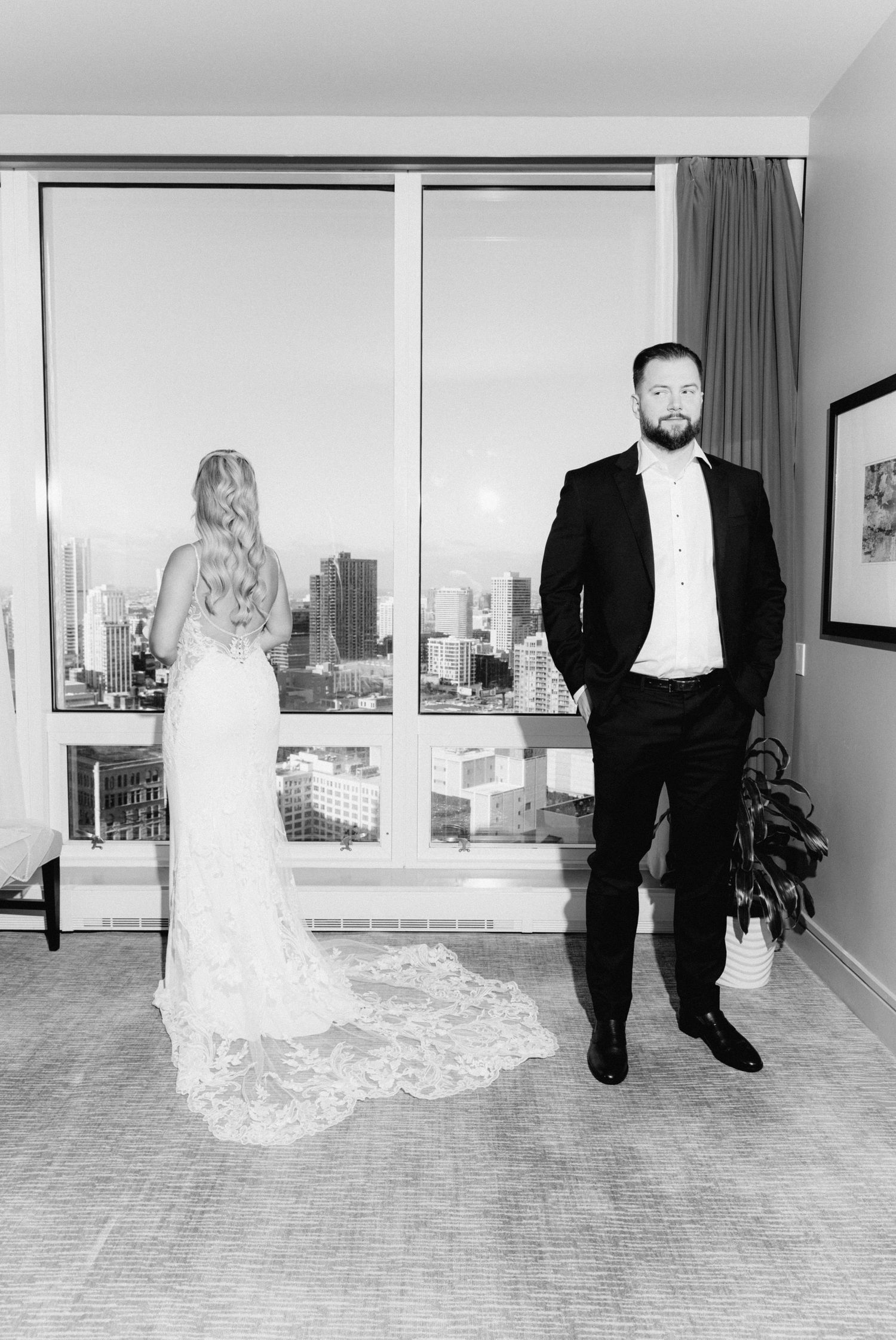 Black and white photo of bride in lace gown facing window as groom in tuxedo stands beside her.