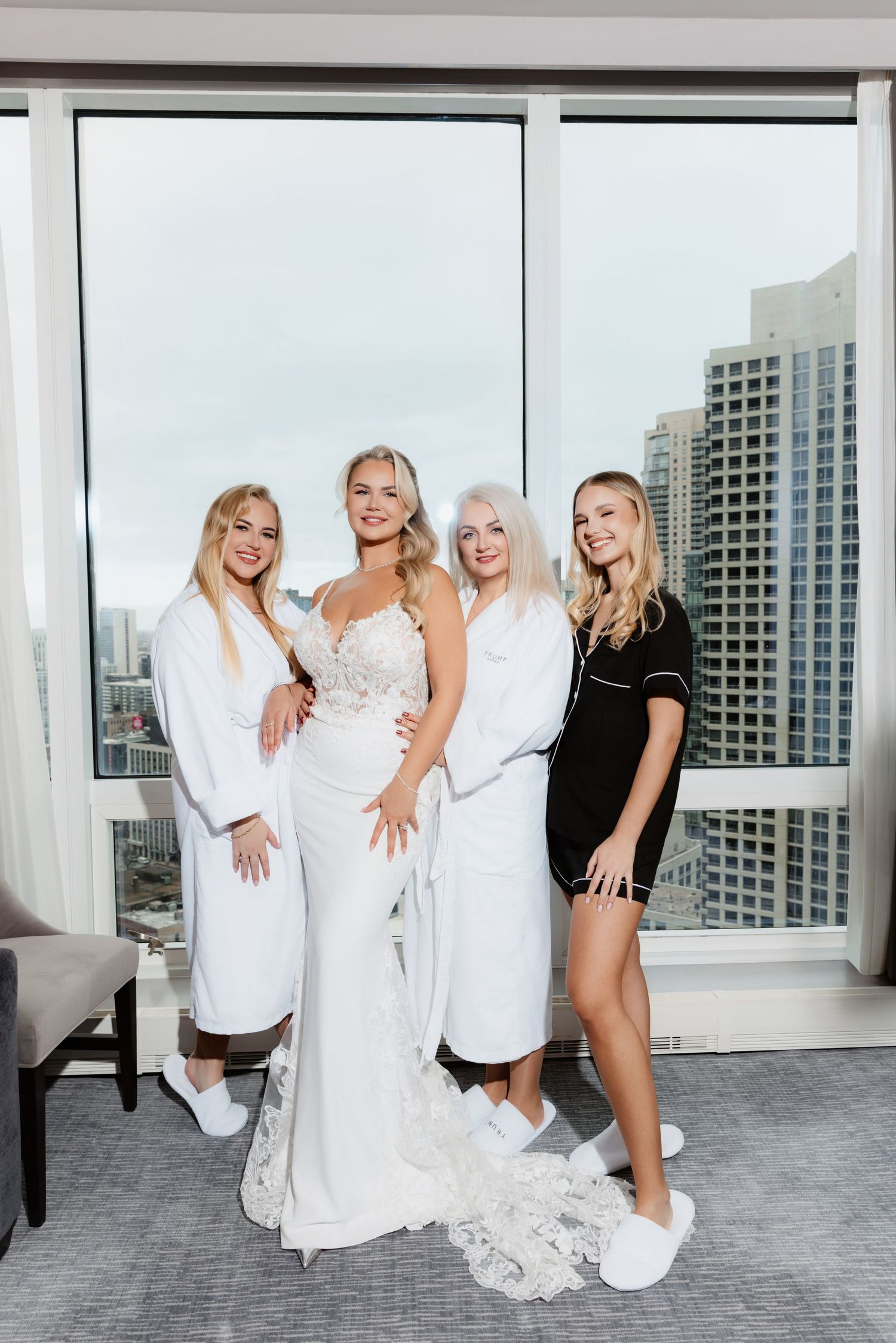 Four blonde women pose in a luxury hotel suite, one in a bridal gown with lace details, others in white robes and black outfit.