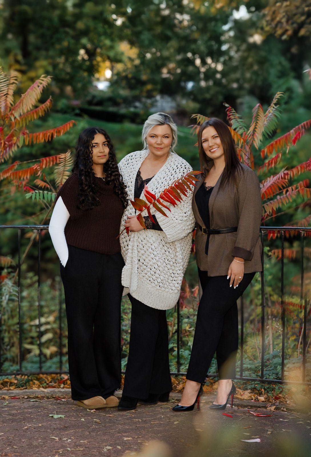 Three women posing together outdoors in autumn park setting with colorful foliage backdrop.