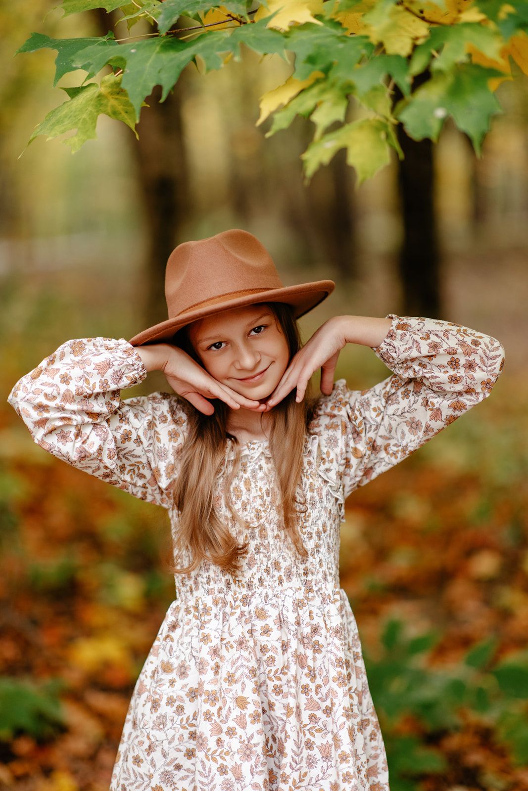 Smiling girl in floral dress and tan hat poses with hands on cheeks under maple tree in fall park.