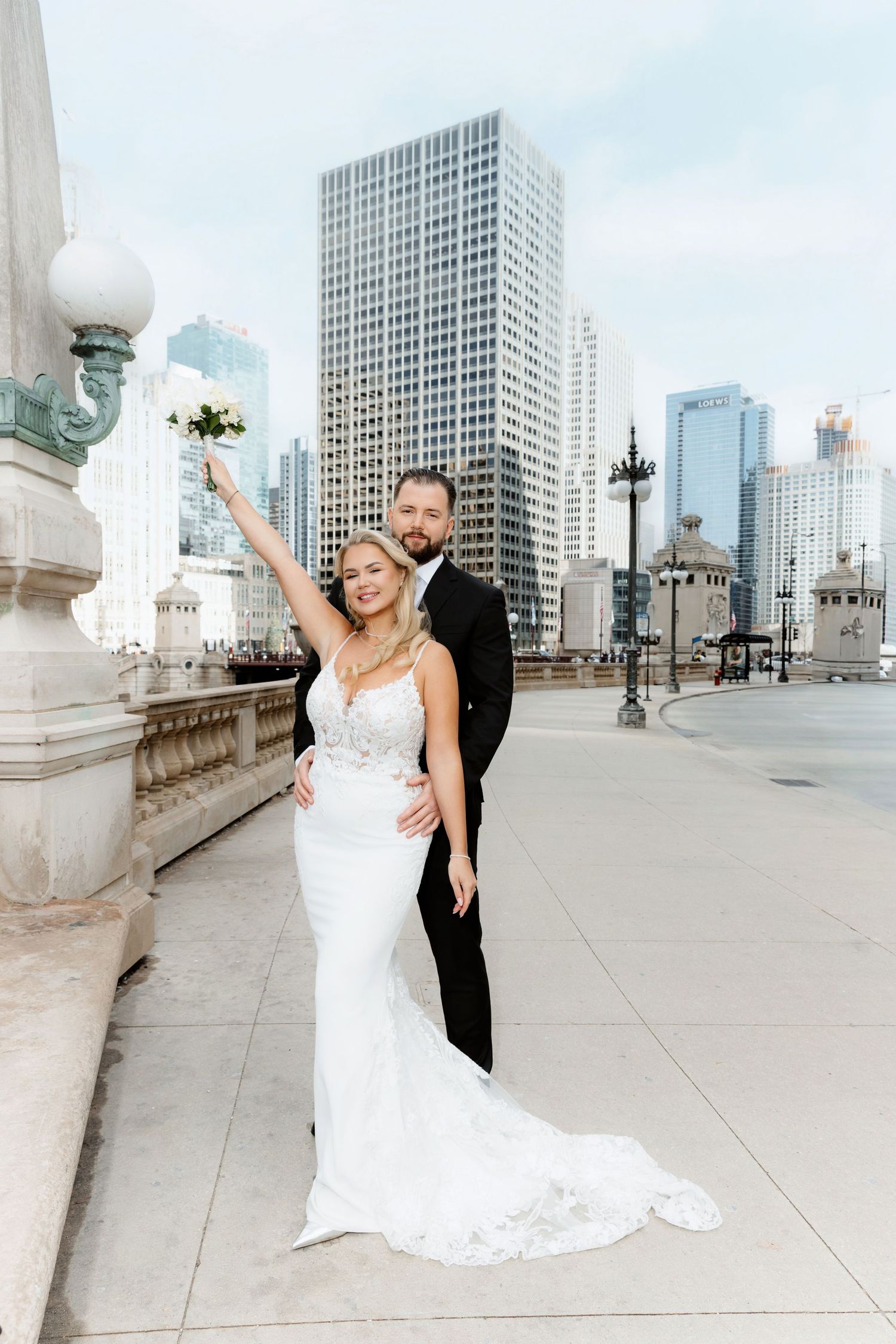 Celebrating bride raising bouquet with groom on a Chicago bridge, tall skyscrapers in background.