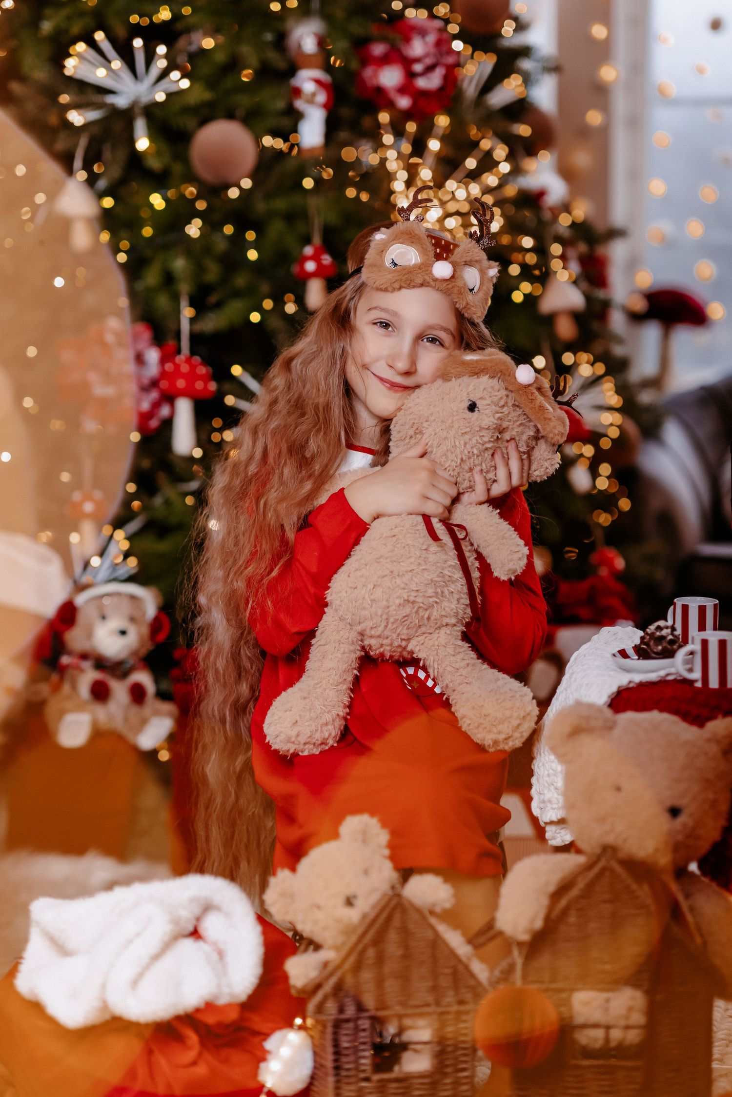 A smiling girl in a red reindeer outfit hugs a plush toy bear in front of a decorated Christmas tree.