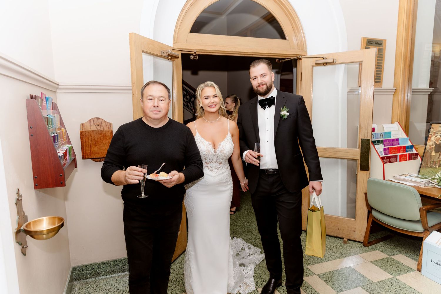 Bride in silver gown and groom in tuxedo pose with a guest at a church entrance.