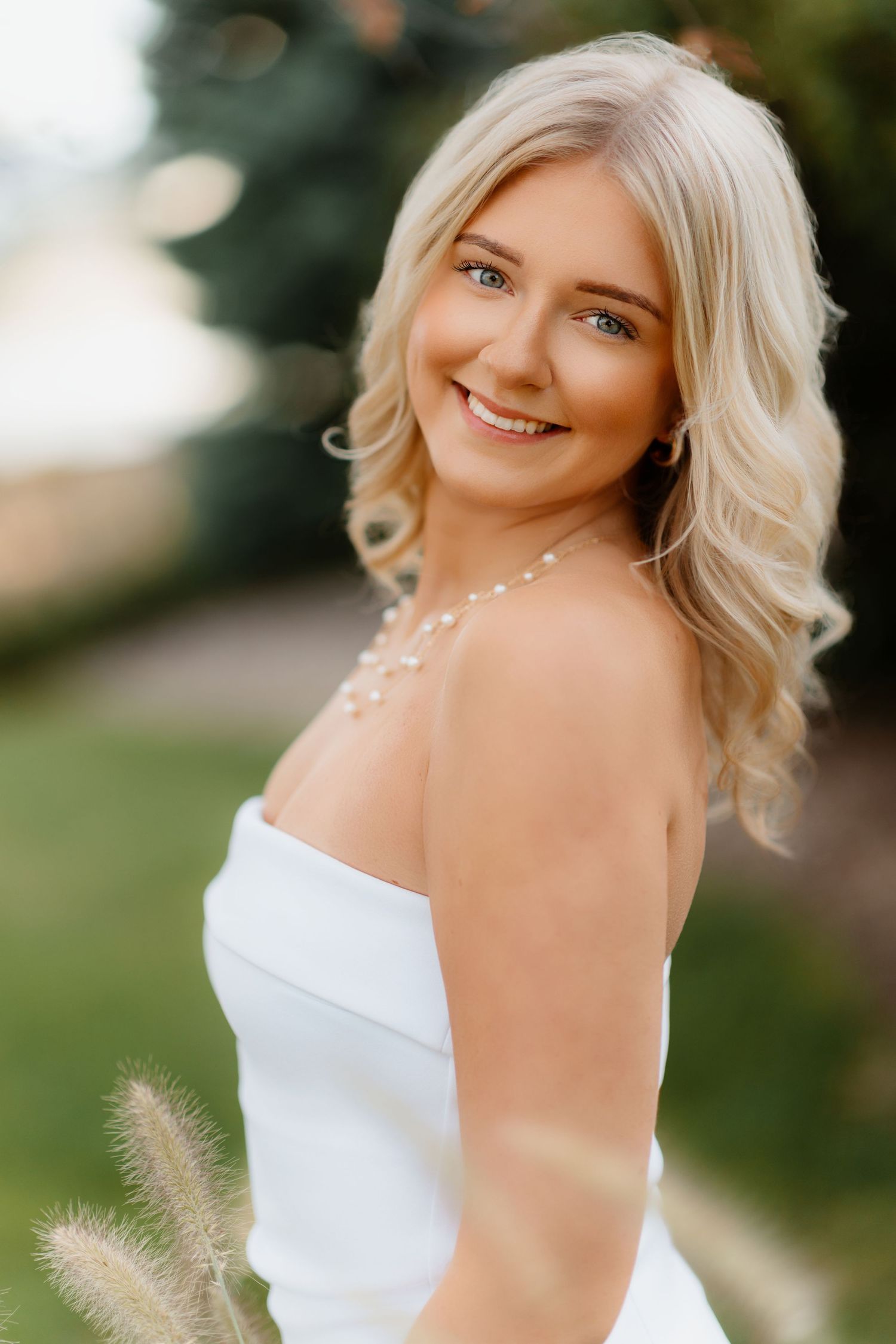 Beautiful blonde woman in white strapless dress smiling outdoors with bokeh background.