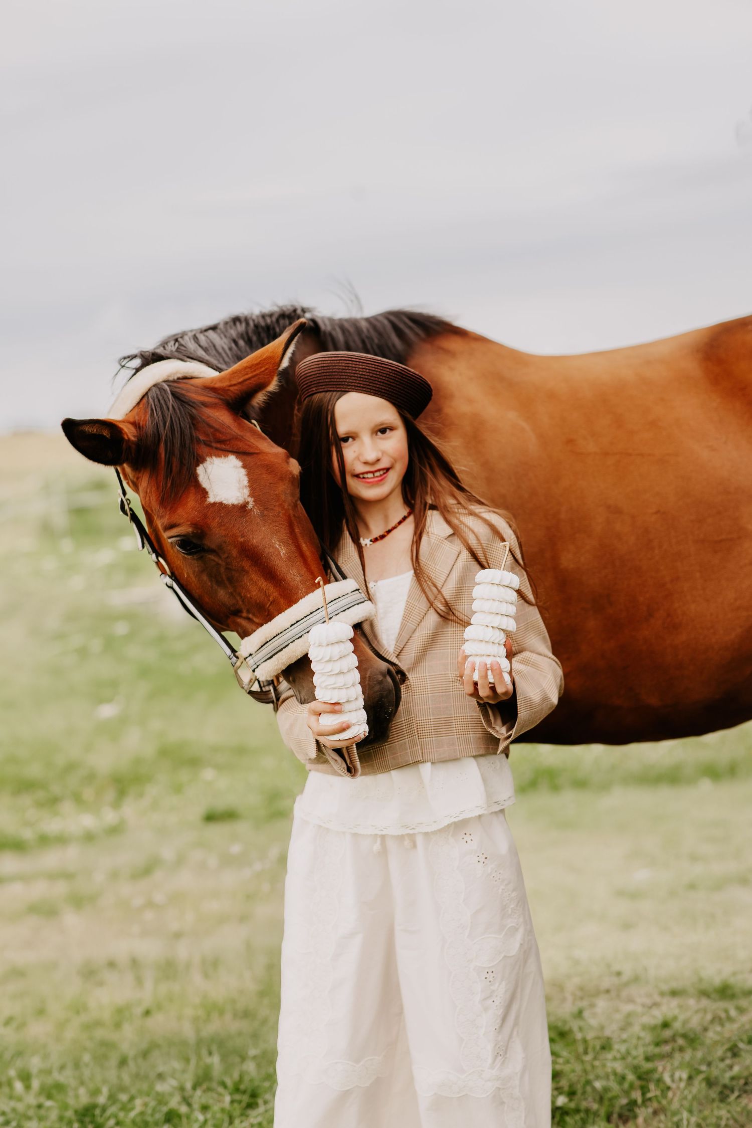 Smiling girl in brown beret and beige blazer posing with a chestnut horse while holding white stacked treats.