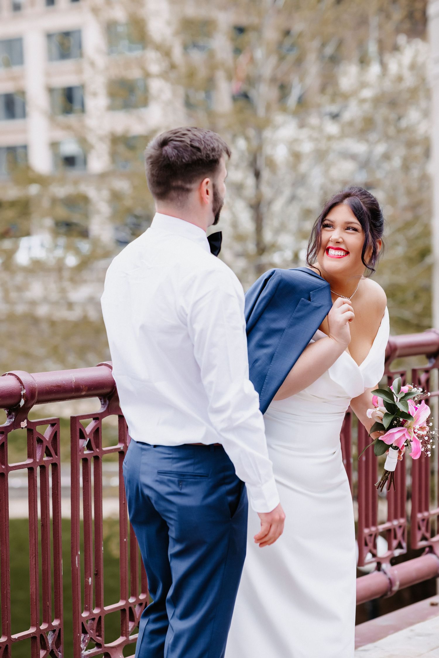 Smiling bride in white gown drapes grooms blue jacket over shoulder while standing on decorative urban bridge.