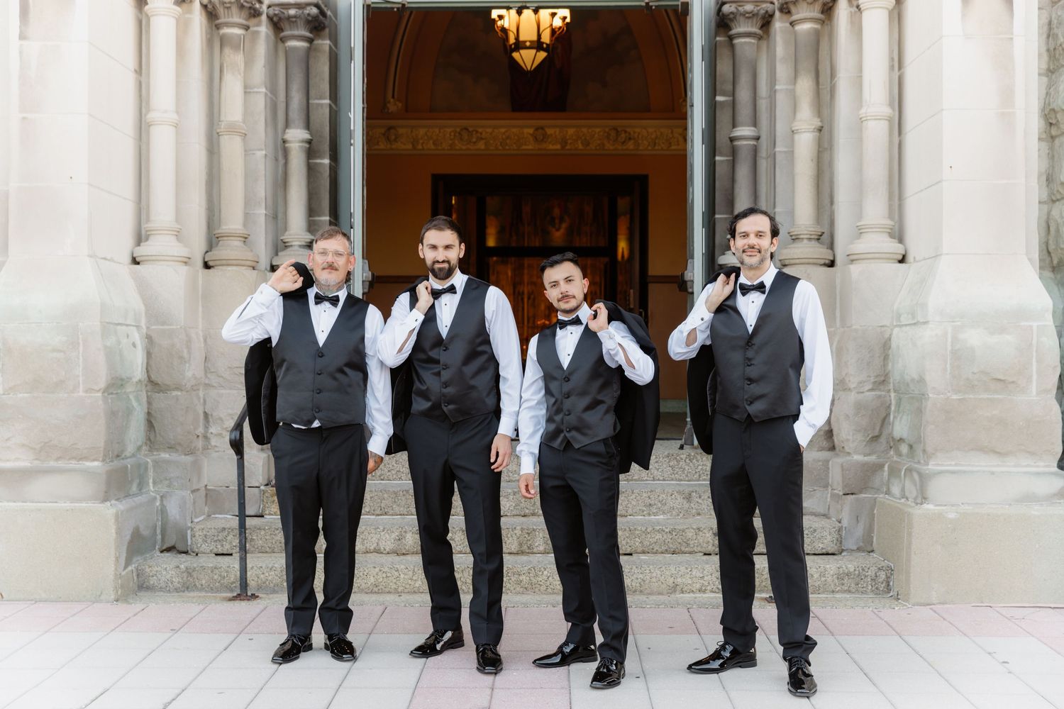 Four groomsmen in black vests and bow ties posing playfully on steps of stone building.