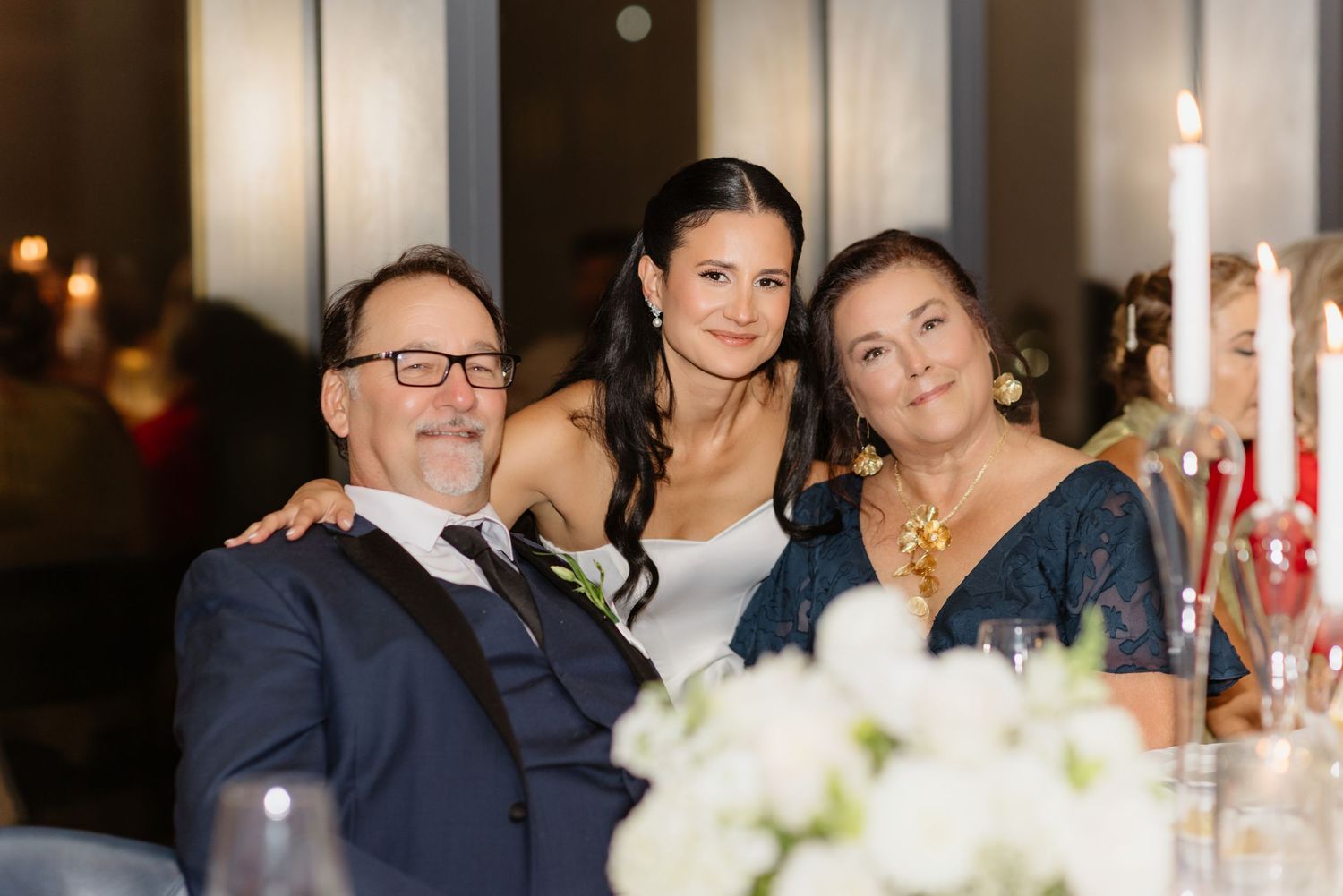 Bride posing with two guests at wedding reception table with white flowers and candlelight.