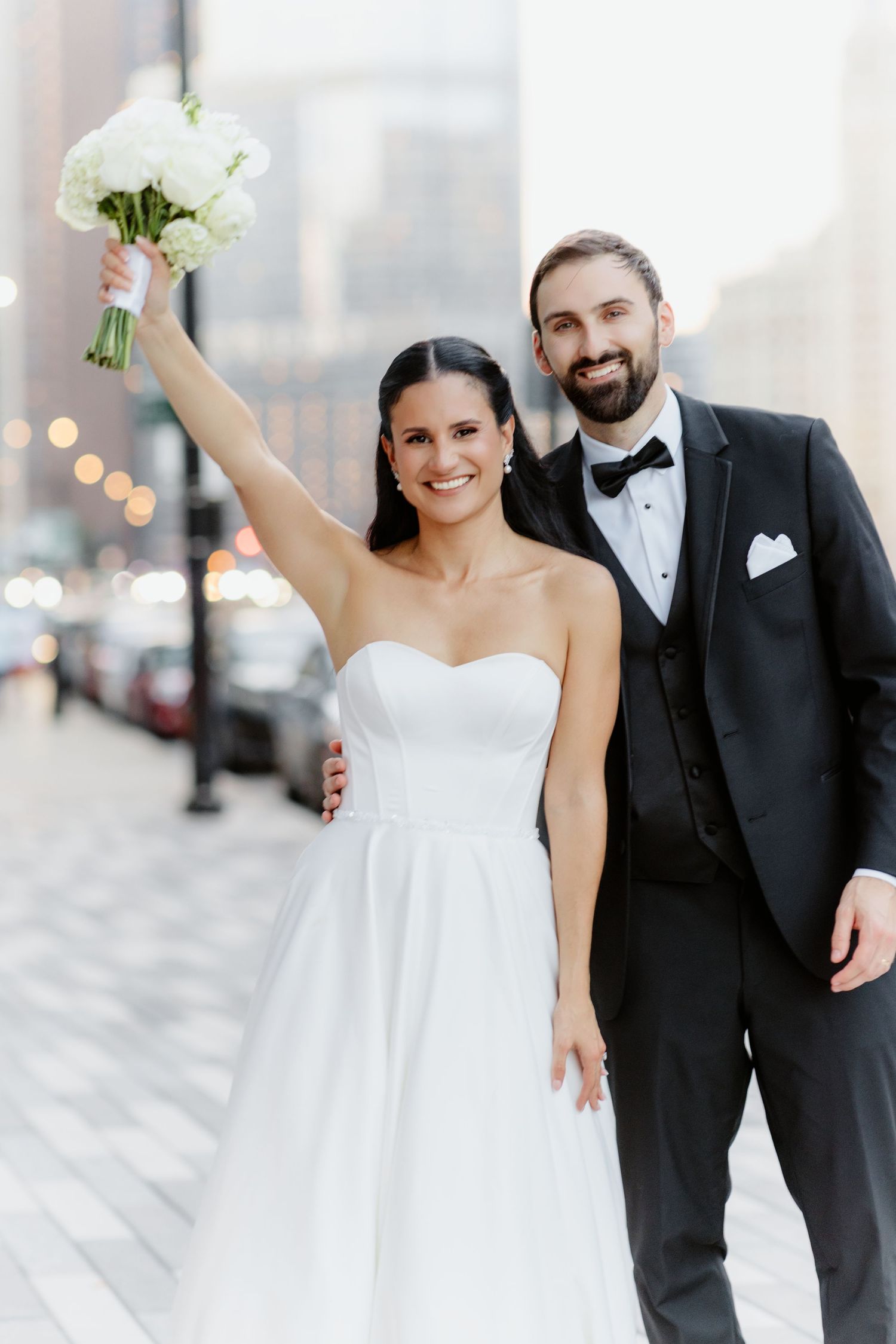 Bride raises white bouquet triumphantly while bearded groom in black tuxedo smiles on a city street.
