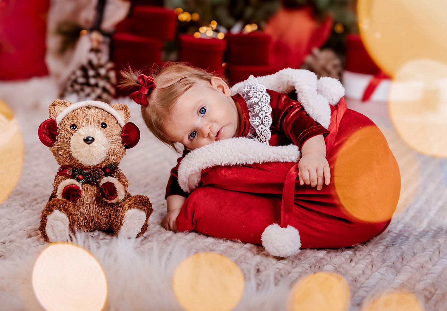 Baby in red Santa outfit lying next to a teddy bear with Christmas tree and gifts in background.