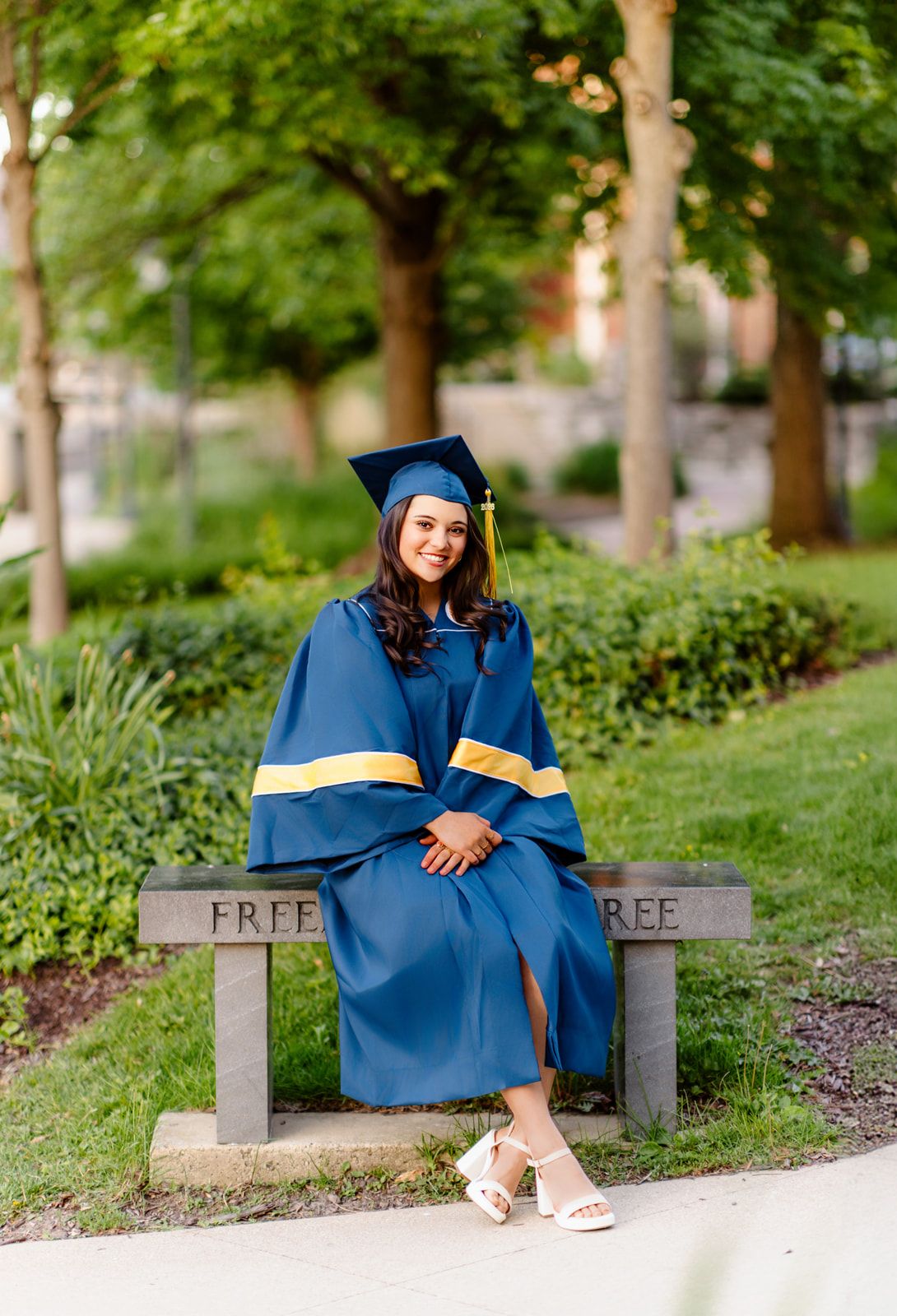Female graduate in navy blue cap and gown sitting on a bench in a lush green park.