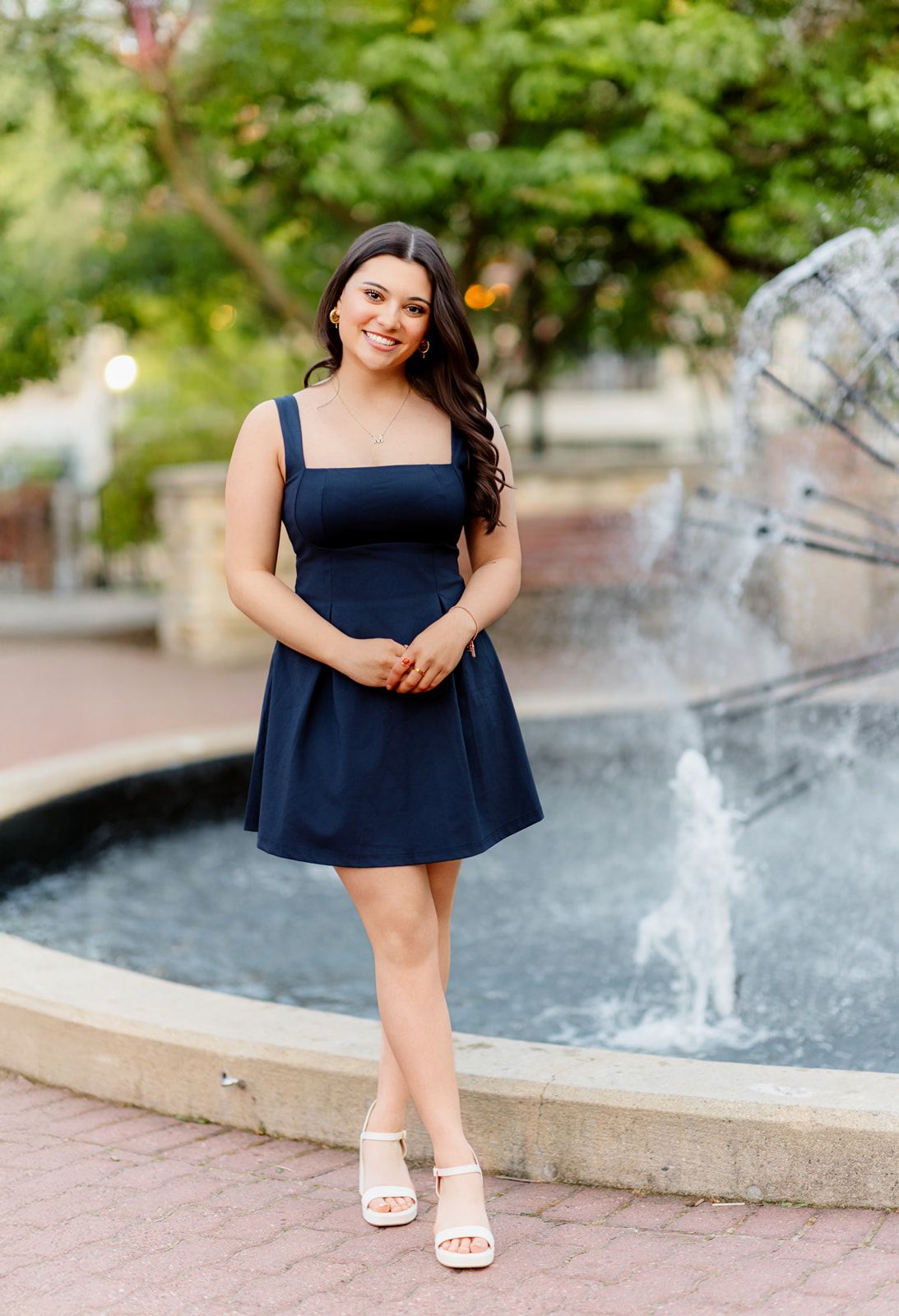 Smiling young woman in navy dress standing near a decorative outdoor fountain.