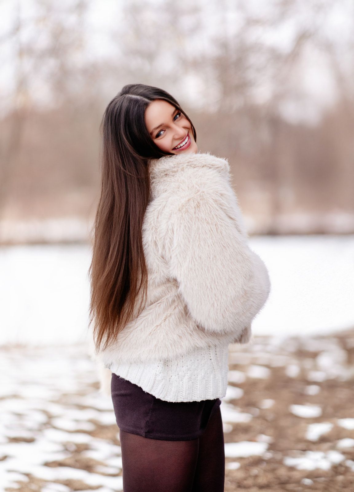 Happy young brunette woman in white fluffy jacket and dark mini skirt smiling over shoulder in snowy winter setting.