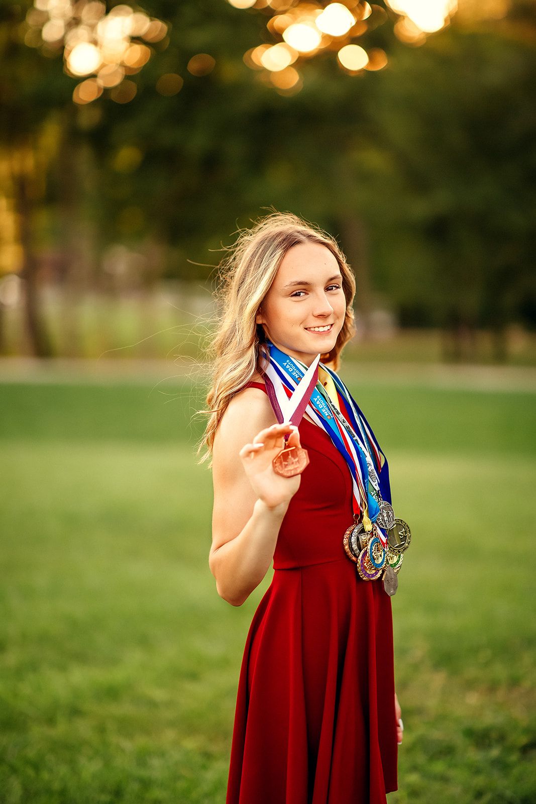 Smiling blonde girl in red dress holding out medals in a sunlit green field.