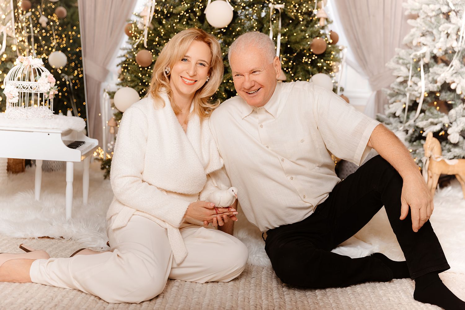 Smiling senior couple in white outfits sitting on floor by decorated Christmas tree in festive living room.