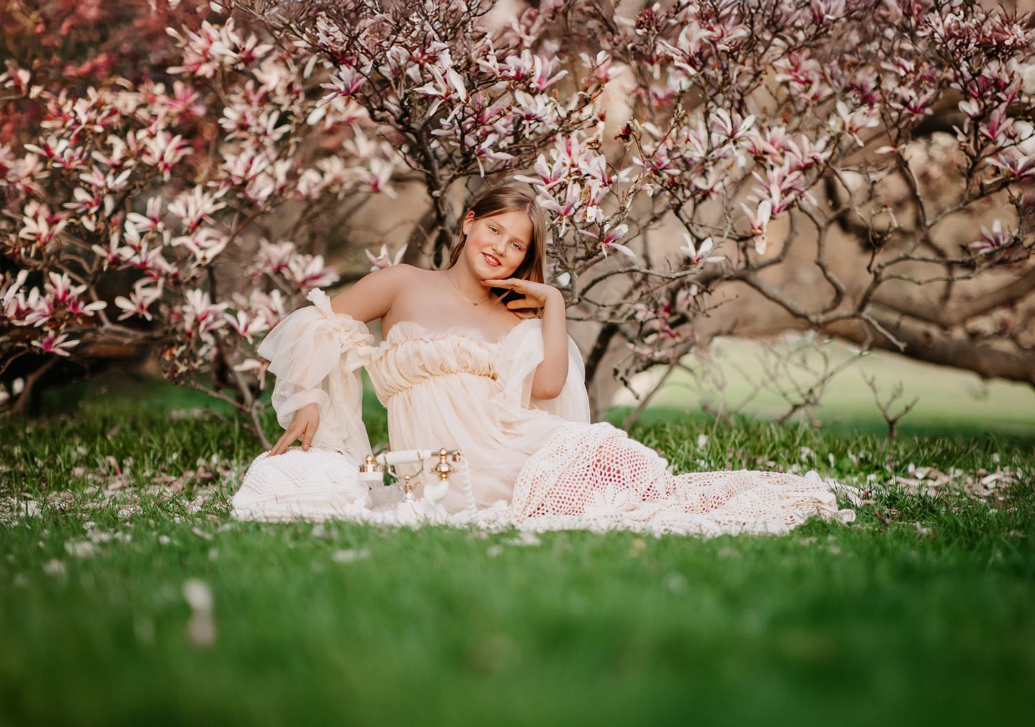 Woman in white dress sitting under blooming magnolia trees on green grass in spring garden.