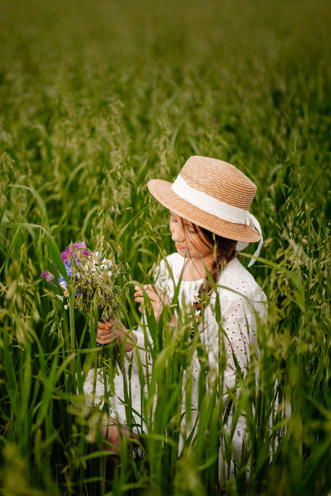 Young girl in straw hat and white dress picking wildflowers in a lush green field.