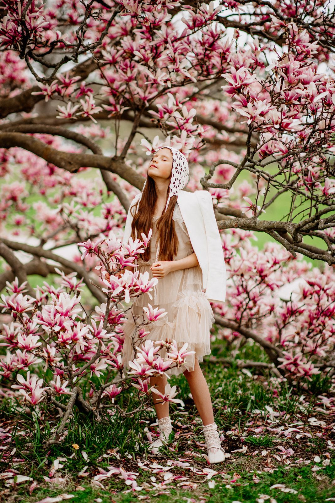 Girl in cream dress and lace headscarf stands among blooming pink magnolia tree branches outdoors.