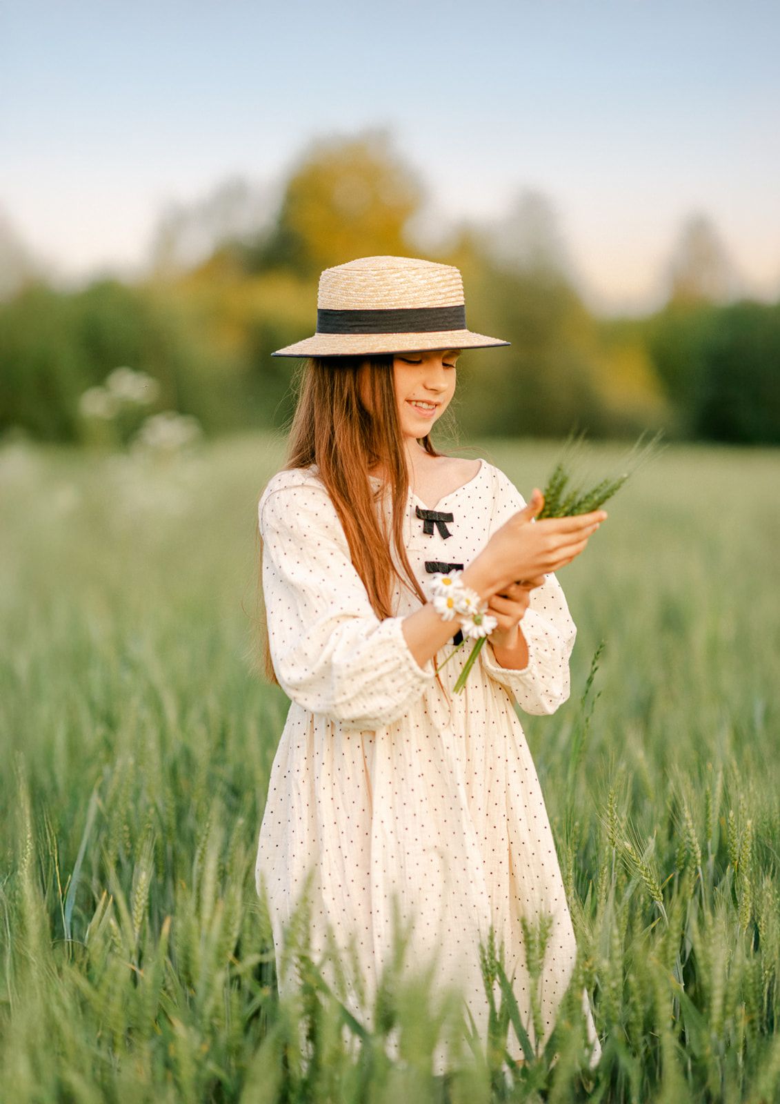 Smiling girl in straw hat and white dress examining wildflowers in a sunny green meadow.