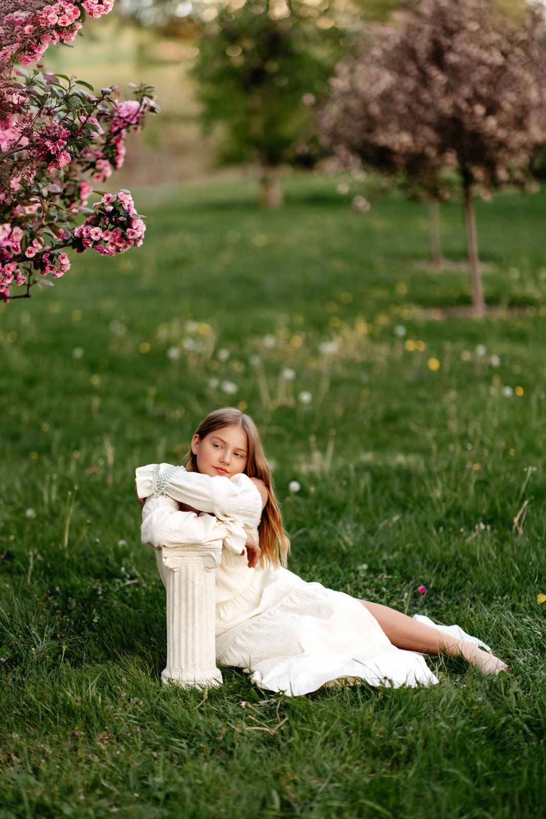 Young girl in white dress sitting on grass leaning against column near blooming pink flowers in spring garden.