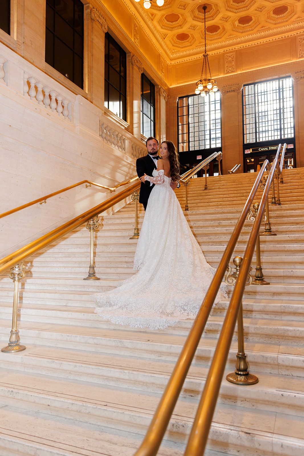 Bride and groom pose on grand marble staircase with gold railings in elegant wedding venue.