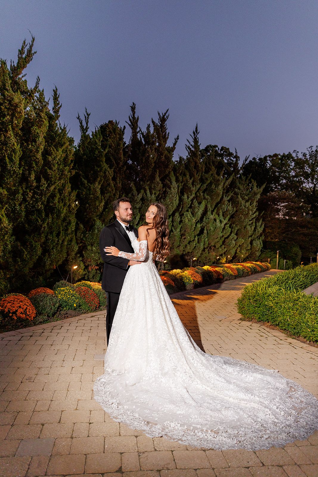 Bride and groom embrace on a garden pathway surrounded by tall evergreens and colorful flowers at dusk.