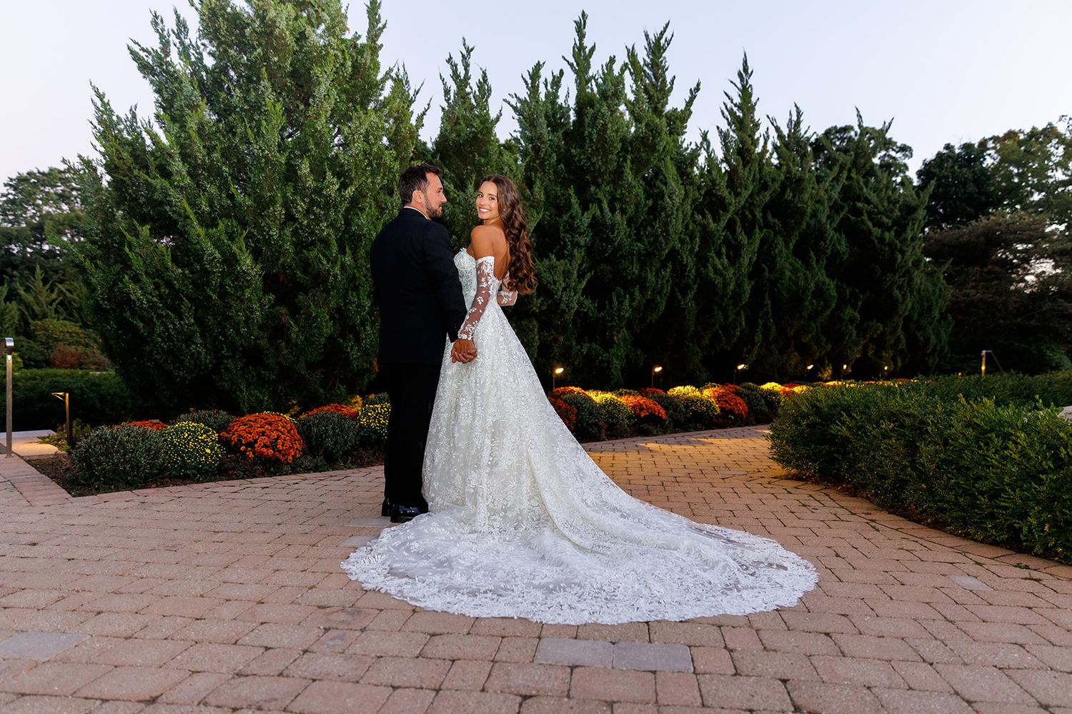 Bride in lace gown with long train stands with groom in garden at twilight.