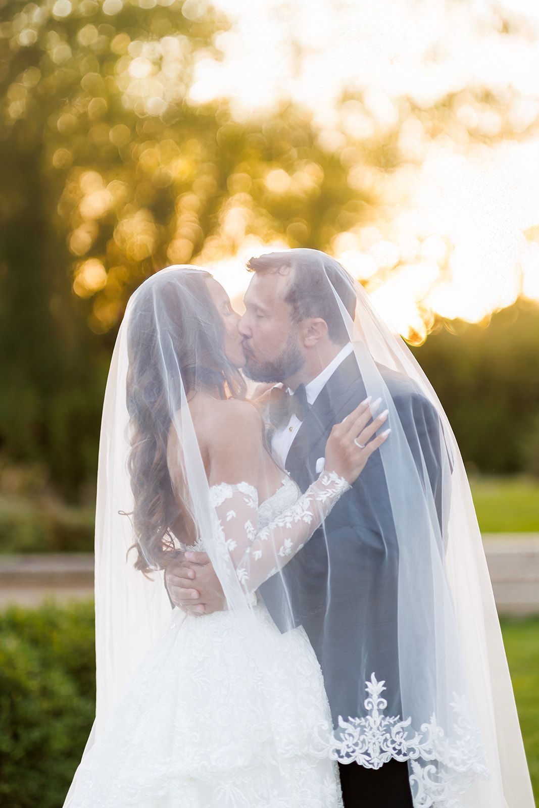 Bride and groom sharing a romantic kiss under a lace-trimmed veil at golden hour outdoors.