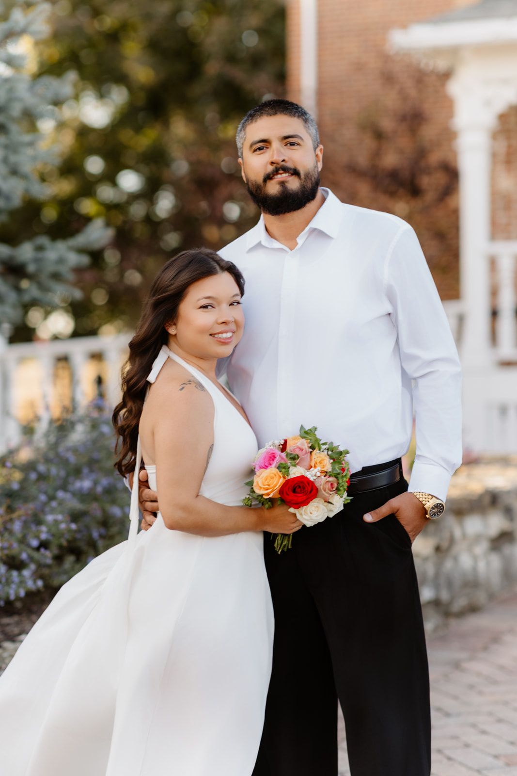 Bride in white dress holding colorful bouquet poses with groom in white shirt and black pants outdoors.